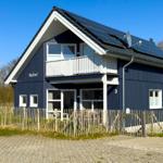Blau gestrichenes Ferienhaus mit Balkon und Solaranlage. Umzäunt mit Holzzaun und Spielplatz.