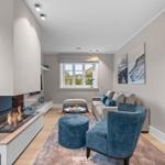 Wohnzimmer mit Kamin, Sofa und blauer Sitzgelegenheit. Fenster mit Blick nach draußen.