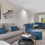 Wohn- und Essbereich mit Sofa, Sesseln und Tischen. Wandbilder und Fenster sichtbar.