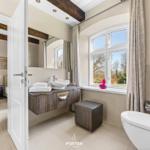 Badezimmer mit Waschbecken, Toilette und Fenster. Schlafzimmer mit Bett und Wandbild sichtbar.
