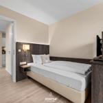 Schlafzimmer mit Bett, Nachttisch und Fernseher. Durchgang zur Küche und Esszone.