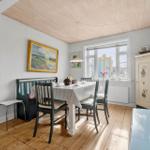 Dining room with table, chairs, and wooden floor. Window, paintings, and cabinet are visible.