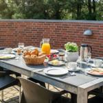 Outdoor area with table, chairs, and breakfast. Background: brick wall and trees.