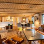 Living area with wooden walls, sofa, chairs, and dining table. Kitchen area in the background.