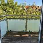 Terrasse mit Holzboden und Glasgeländer. Blick auf grüne Bäume und rote Dächer.