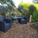 Garten mit zwei Sesseln, Tisch und Wein. Holzmulch, Pflanzen und Steinmauer.