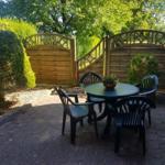 Terrasse mit Tisch und Stühlen, Holztor und Beete im Hintergrund.