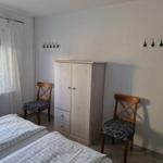 Zimmer mit Doppelbett, Kleiderschrank und zwei Stühlen. Fenster mit Vorhängen.