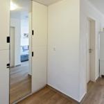 Flur mit Spiegeltüren, weißen Wänden und Holzboden. Blick in Wohnzimmer mit Sofa.