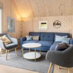 Wohnzimmer mit blauem Sofa, zwei Sesseln und runden Tisch. Holzwände und große Fenster.