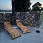 Zwei Holzliegestühle stehen auf einer gepflasterten Terrasse vor einer Steinmauer mit Beleuchtung.