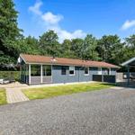 Ein graues Ferienhaus mit Terrasse, Liegestühlen und Grillplatz. Grün umgeben mit Wald.