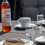 Außenbereich mit Tisch, Tassen, Gläsern und Gebäck. Eine Flasche mit Getränk steht daneben.