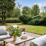 Terrasse mit Sitzgruppe, Tisch und Pflanze. Grünfläche und Bäume im Hintergrund.