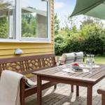 Terrasse mit Tisch, Bänken und Sonnenschirm. Hintergrund: grüner Garten und gelbes Haus.