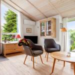 Wohnraum mit Kamin, zwei Sesseln und Holztisch. Große Fenster zeigen grüne Bäume.