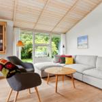 Wohnzimmer mit Sofa, Sessel und Holzdecke. Fenster blicken auf grüne Außenanlage.