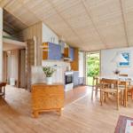 Wohn- und Essbereich mit Küche unter Holzdecke. Tisch mit Stühlen, Sofa und Pflanze.