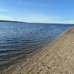 Strand mit Sand und Steinen am Wasser, Boot im Hintergrund unter blauem Himmel.