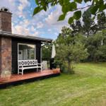 Schwarzes Holzhaus mit Stein-Säule und Terrasse. Grünfläche und Bäume im Hintergrund.