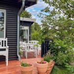 Terrasse mit weißem Tisch und Stühlen, Pflanzen und Obstbaum. Schwarzes Holzhaus mit Fenster.