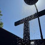 Duschkopf mit Wasserstrahl vor blauem Himmel und Holzstruktur