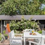 Hinterhof mit weißem Tisch, Stühlen und Blumenhecken. Schwarzes Holzhaus im Hintergrund.