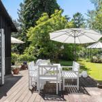 Terrasse mit weißem Tisch und Stühlen unter Sonnenschirm. Grünflächen und Gewächshaus im Hintergrund.