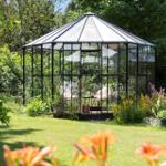 Glasveranda mit Sitzgelegenheit im Garten, umgeben von Pflanzen und Blumen.