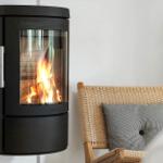 Wandmontierte Kaminofen mit brennendem Feuer neben einem geflochtenen Stuhl mit Kissen.