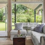 Ein Wohnbereich mit Sofa, Kissen und Tisch. Durchgehende Glasfront zeigt Garten.
