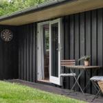 Schwarzer Holzschuppen mit Terrasse, Tisch, Stühlen und Dartscheibe an der Wand.
