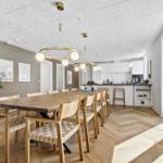 Esszimmer mit langem Holztisch und Stühlen. Küche im Hintergrund. Holzboden und große Fenster.