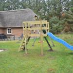 Hinterhof mit Holzspielplatz, blauer Rutsche und Sandkasten. Im Hintergrund ein Haus mit Strohdach.