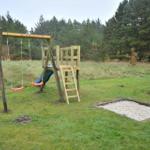 Holzspielplatz mit Schaukel, Rutsche und Sandkasten auf grünem Rasen.