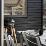 Außenbereich mit Grill, Tisch und Stühlen vor einem Haus mit dunkler Holzfassade.