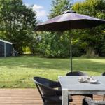 Terrasse mit Tisch, Stühlen und Sonnenschirm. Grünfläche und Holzdecke. Hintergrund mit Gartenhaus und Bäumen.