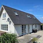 Weißes Haus mit dunklem Dach, Terrasse und Gartenbereich unter blauem Himmel.