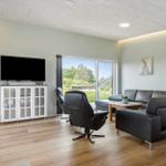 Wohnzimmer mit Sofa, Sessel und Fernseher. Große Fenster mit Blick auf Grünfläche.