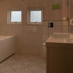 Badezimmer mit Badewanne, Waschbecken und Toilette. Holzwände und Fenster mit Blick auf Wasser.