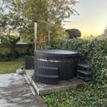 Schwarzer Whirlpool auf Holzterrasse mit Garten und Bäumen im Hintergrund.