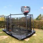 Trampolin mit Korb im Garten, umgeben von Hecke und Gras.