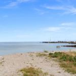 Strand mit Blick auf Hafen und Segelboote unter blauem Himmel.