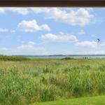 Durch ein Fenster blickt man auf eine Wiese mit Gras und einen See unter blauem Himmel mit Wolken.