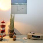 Tisch mit Lautsprecher, Vase, Vogel- und Leuchtturmfiguren sowie Wandbild mit Strandmotiv.