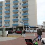 Hochhaus mit Balkonen und Geschäften im Erdgeschoss. Menschen und Fahrräder sind sichtbar.