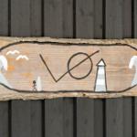 Holzschild mit Strandmotiv und Leuchtturm, an dunkler Holzwand angebracht.