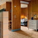 Living area with wooden walls and bedroom in background. White dresser and tiled wall visible.