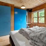 Bedroom with wooden walls, blue door and wardrobe, bed with patterned bedding and window with view of forest.