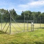 Fenced tennis court with entrance gate and green surroundings.